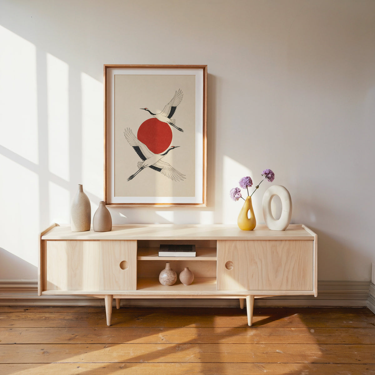 two cranes crossing a red sun print shown in a wood frame with a white mat above a pale wood media console