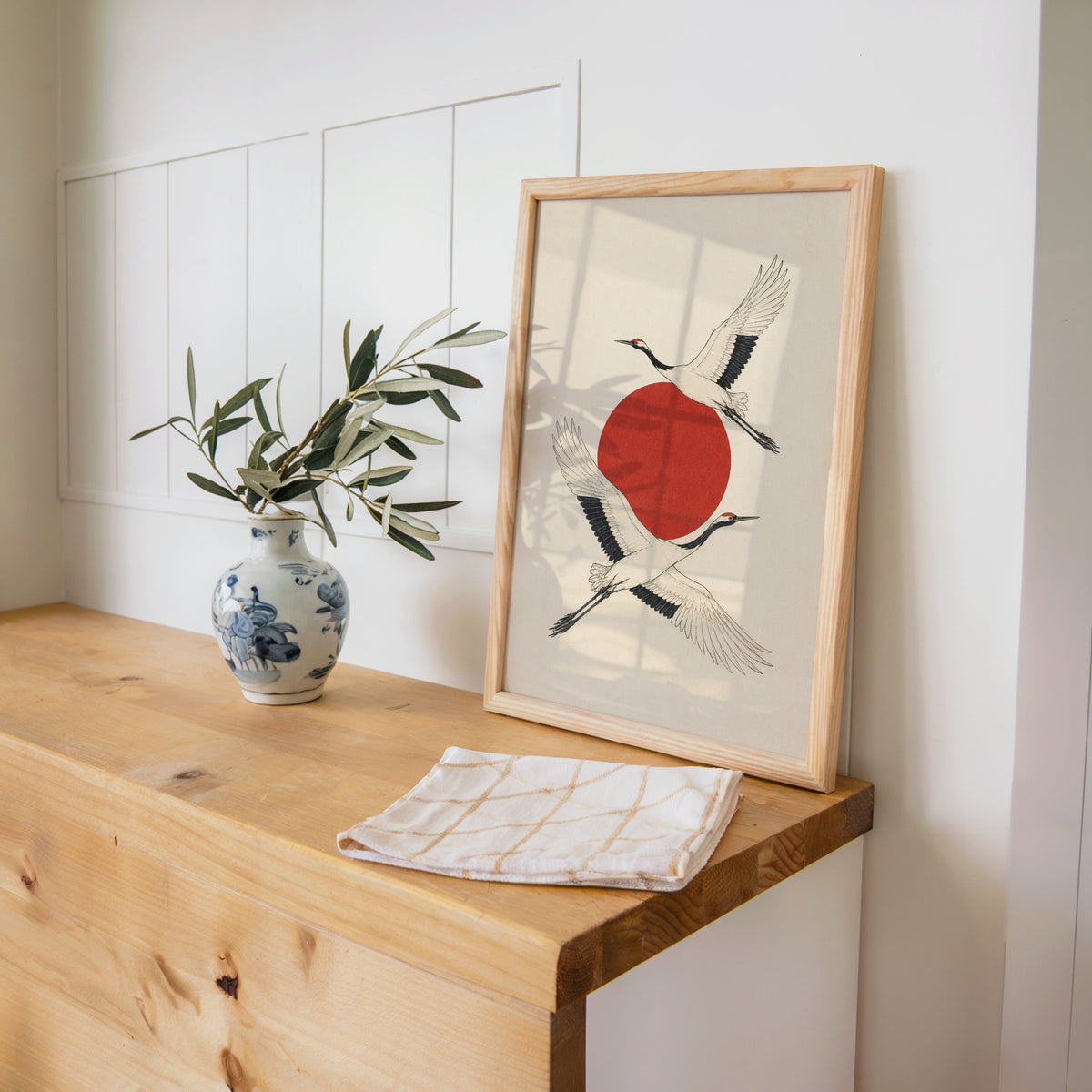 two cranes crossing a red sun print in a pine frame on a wooden counter beside a blue and white porcelain vase