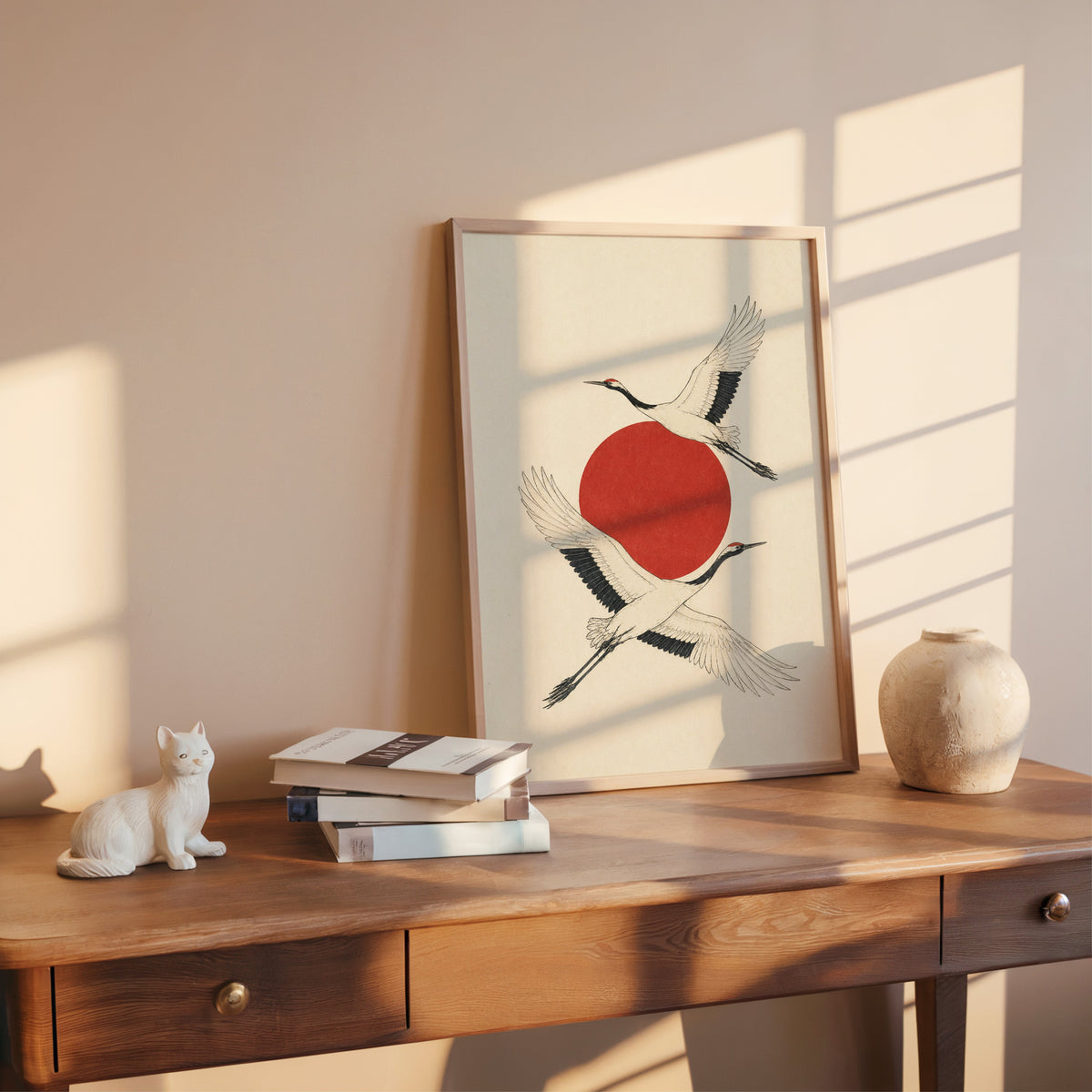 two cranes crossing a red sun print shown in a rose gold frame leaning on a wooden writing desk with books