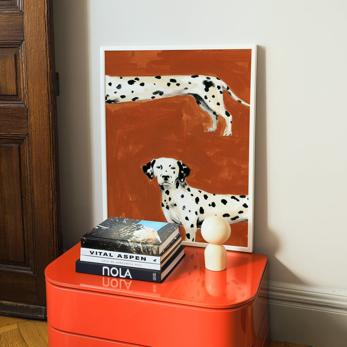 Rust red Dalmatian print in a white frame on a glossy orange cabinet with books