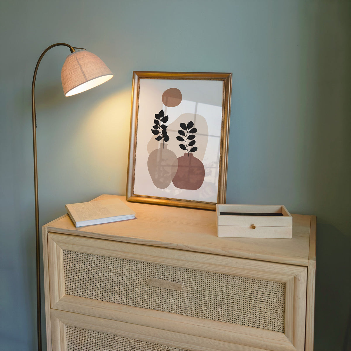 Two vases and terracotta sun abstract botanical print shown in a gold frame on a cane dresser lit by a floor lamp