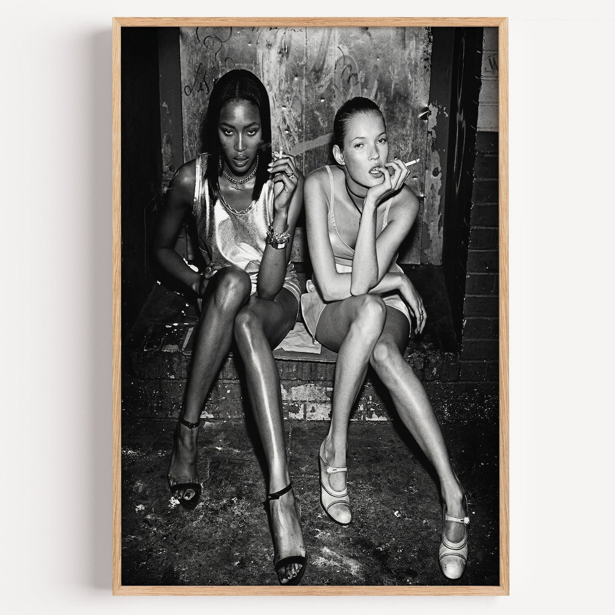 Two women seated on brick doorway steps print in a light oak frame on a plain white wall