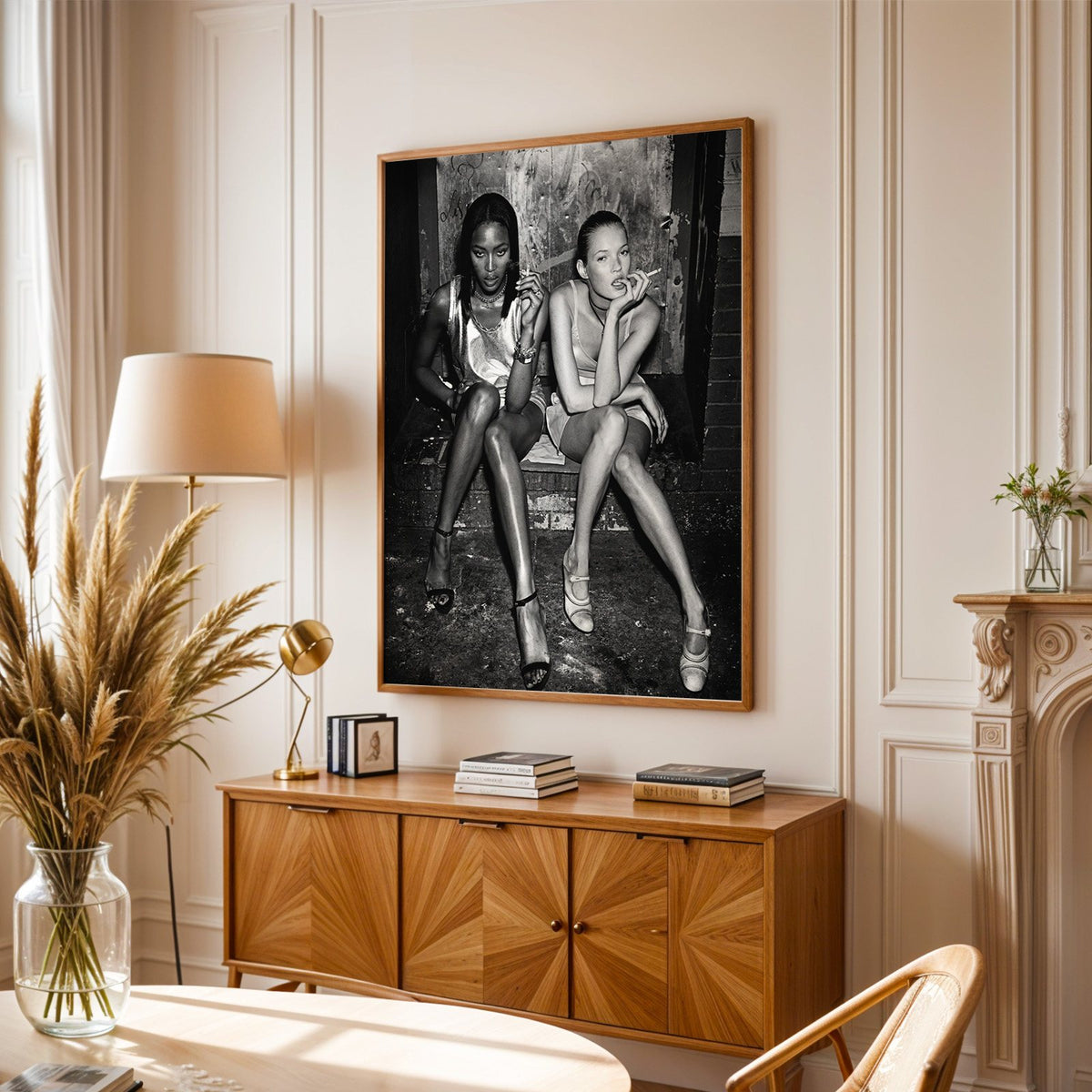 Two women seated on brick doorway steps print in an oak frame above a sunburst wood sideboard with pampas grass