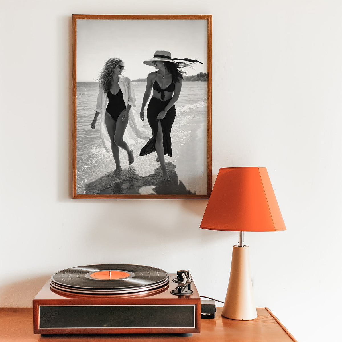 Two women walking the shoreline and laughing print in a walnut frame above a record player and orange lamp
