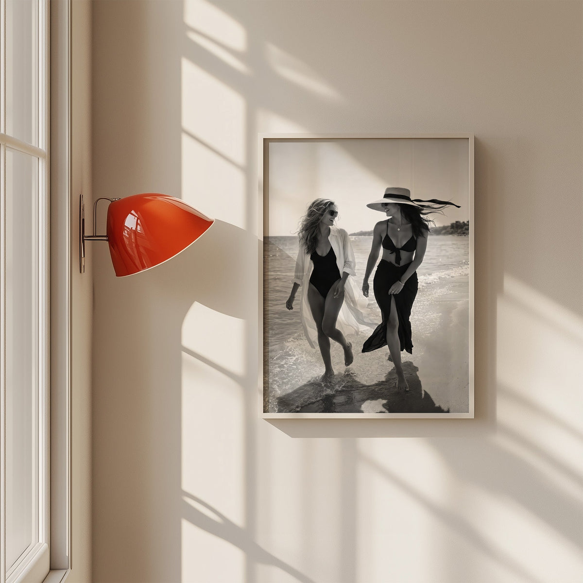 Two women walking the shoreline and laughing print in a white frame on a sunlit wall beside a red sconce