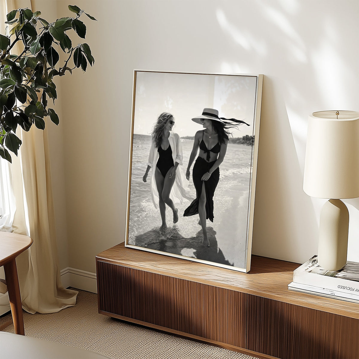Two women walking the shoreline and laughing print in a white frame on a walnut sideboard beside a ceramic lamp
