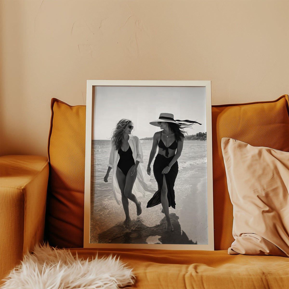 Two women walking the shoreline and laughing print in a white frame leaning on a mustard velvet sofa