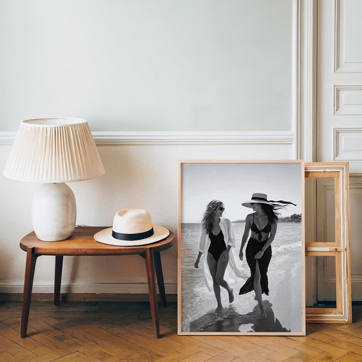 Two women walking the shoreline and laughing print in a pine frame on a wood floor beside a lamp and straw hat