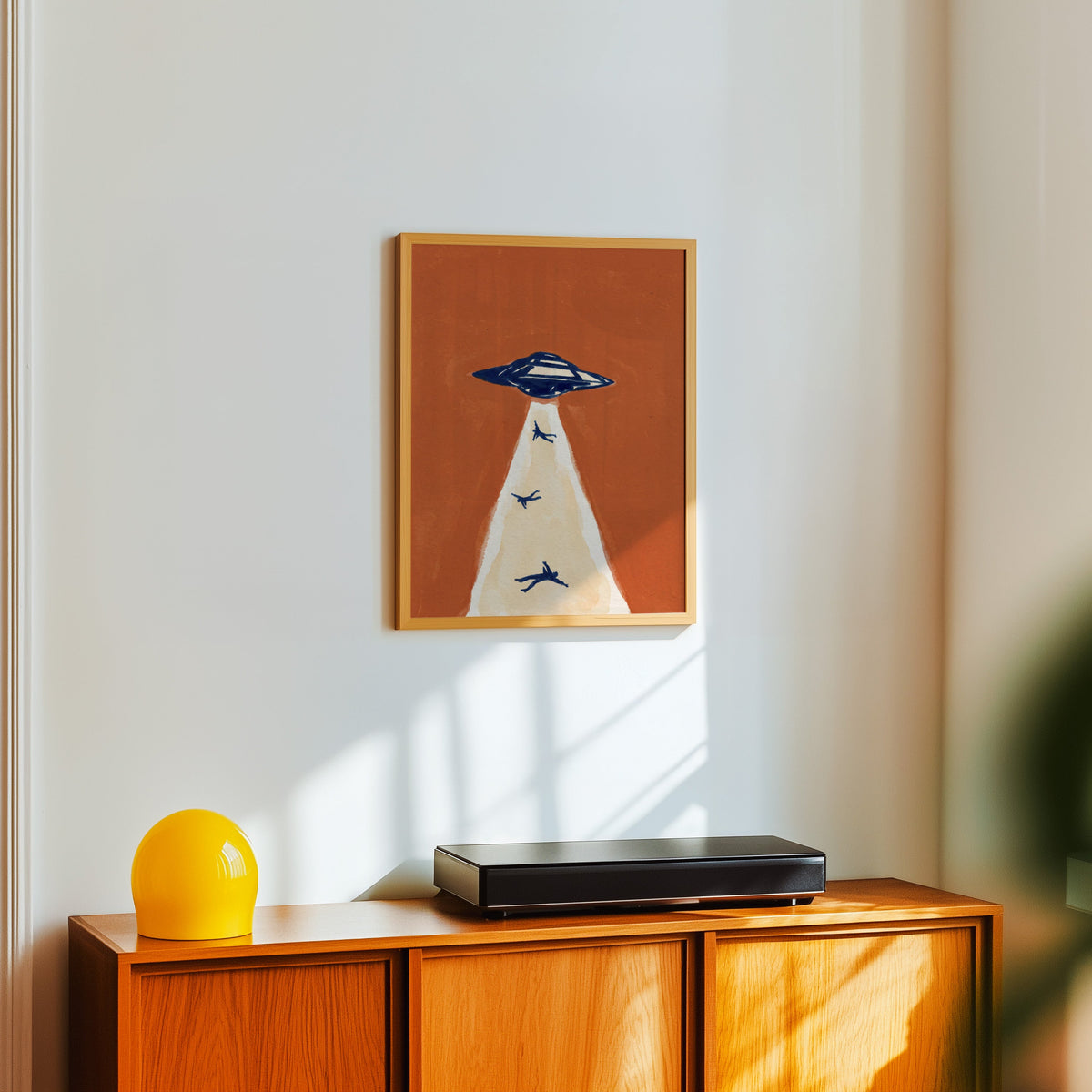 Rust and navy UFO print in a light oak frame above a teak sideboard with a yellow lamp
