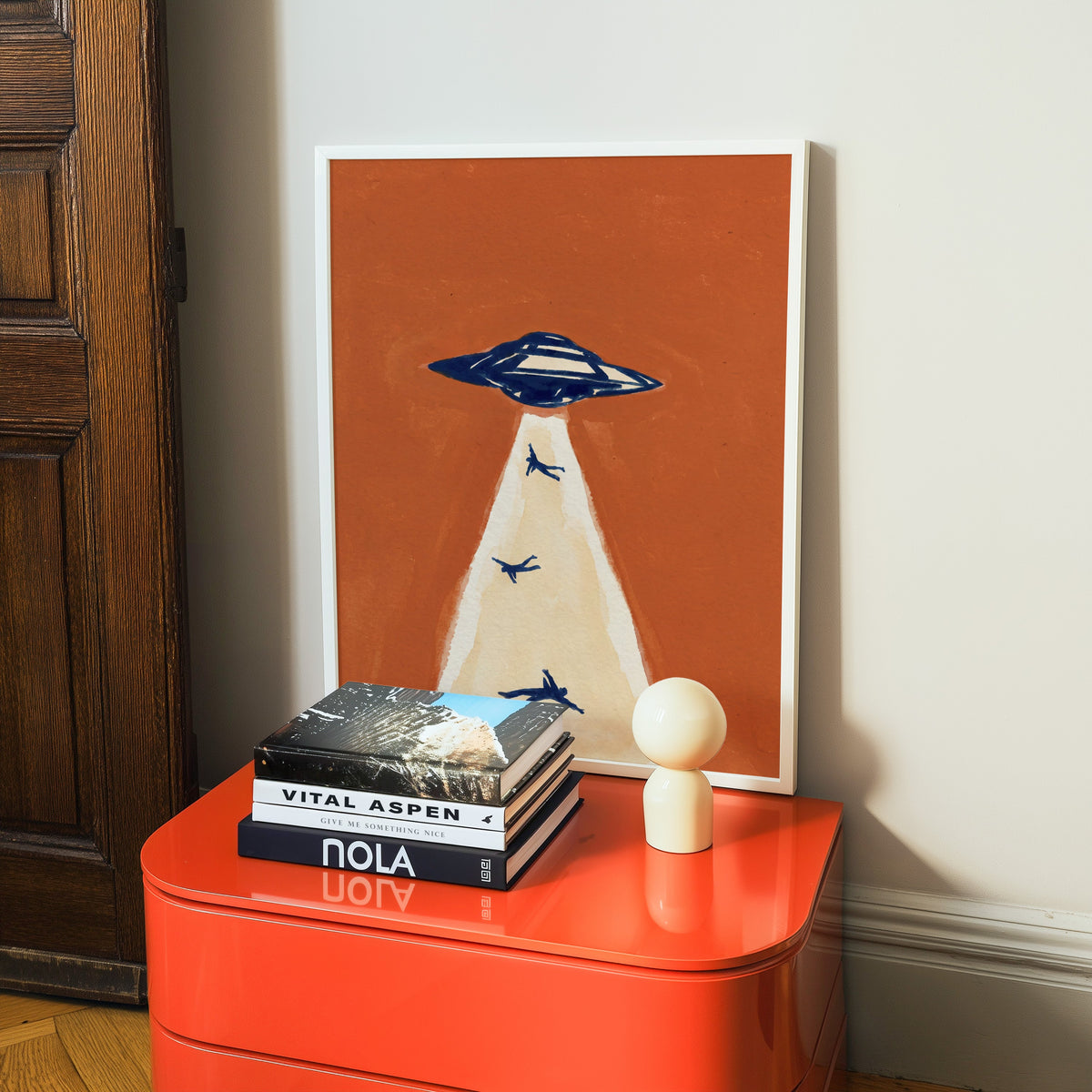 Retro sci-fi print in a white frame on a glossy orange cabinet with books