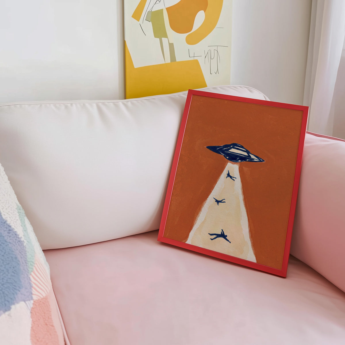 Alien abduction wall art print shown in a red frame leaning on a blush pink sofa