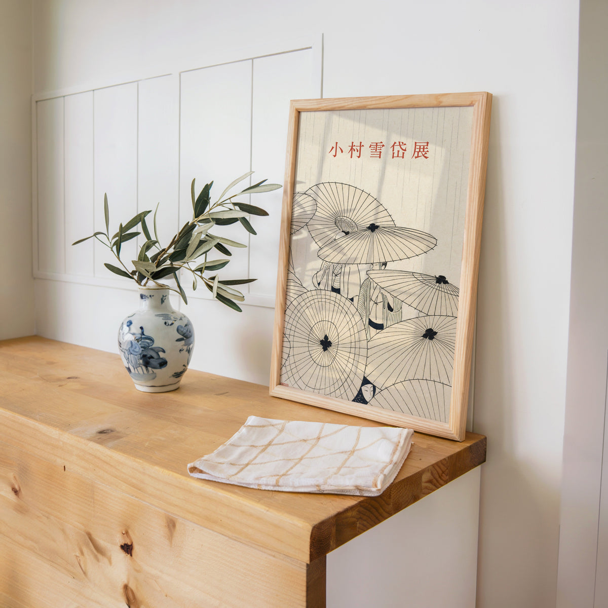 paper umbrellas in the rain line art print in a pine frame on a wooden counter beside a blue and white porcelain vase