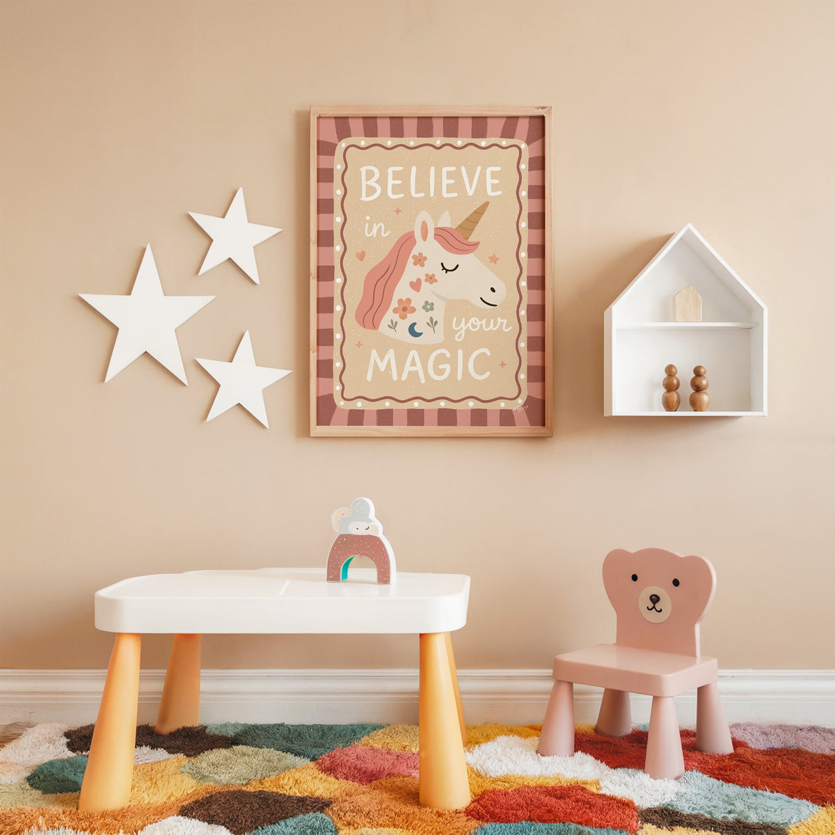 Unicorn head with magic quote nursery print in an oak frame on a peach wall above a kids table with wall stars