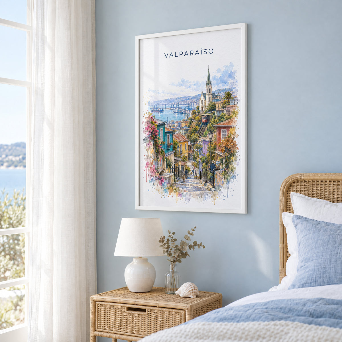 Valparaíso watercolor print in a white frame on a pale blue wall beside a rattan bed in a coastal bedroom