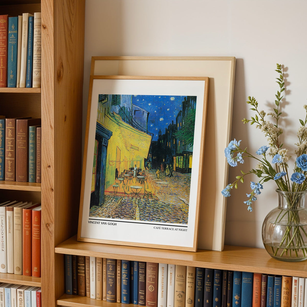 Van Gogh poster in an oak frame leaning on a bookshelf beside blue flowers