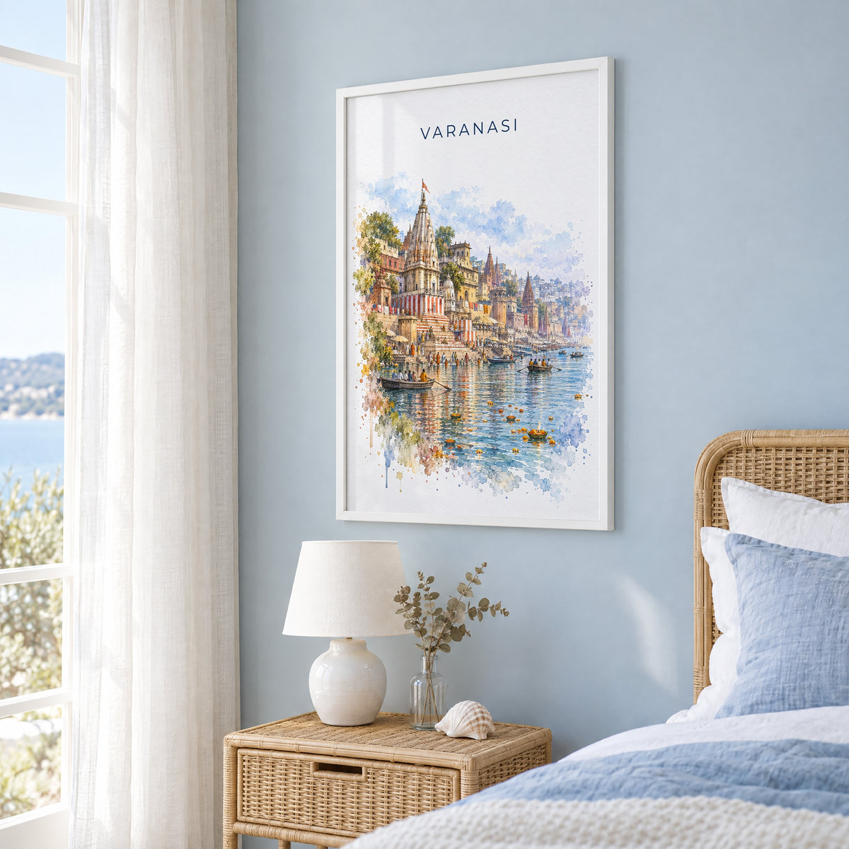 Varanasi watercolor print in a white frame on a pale blue wall beside a rattan bed in a coastal bedroom
