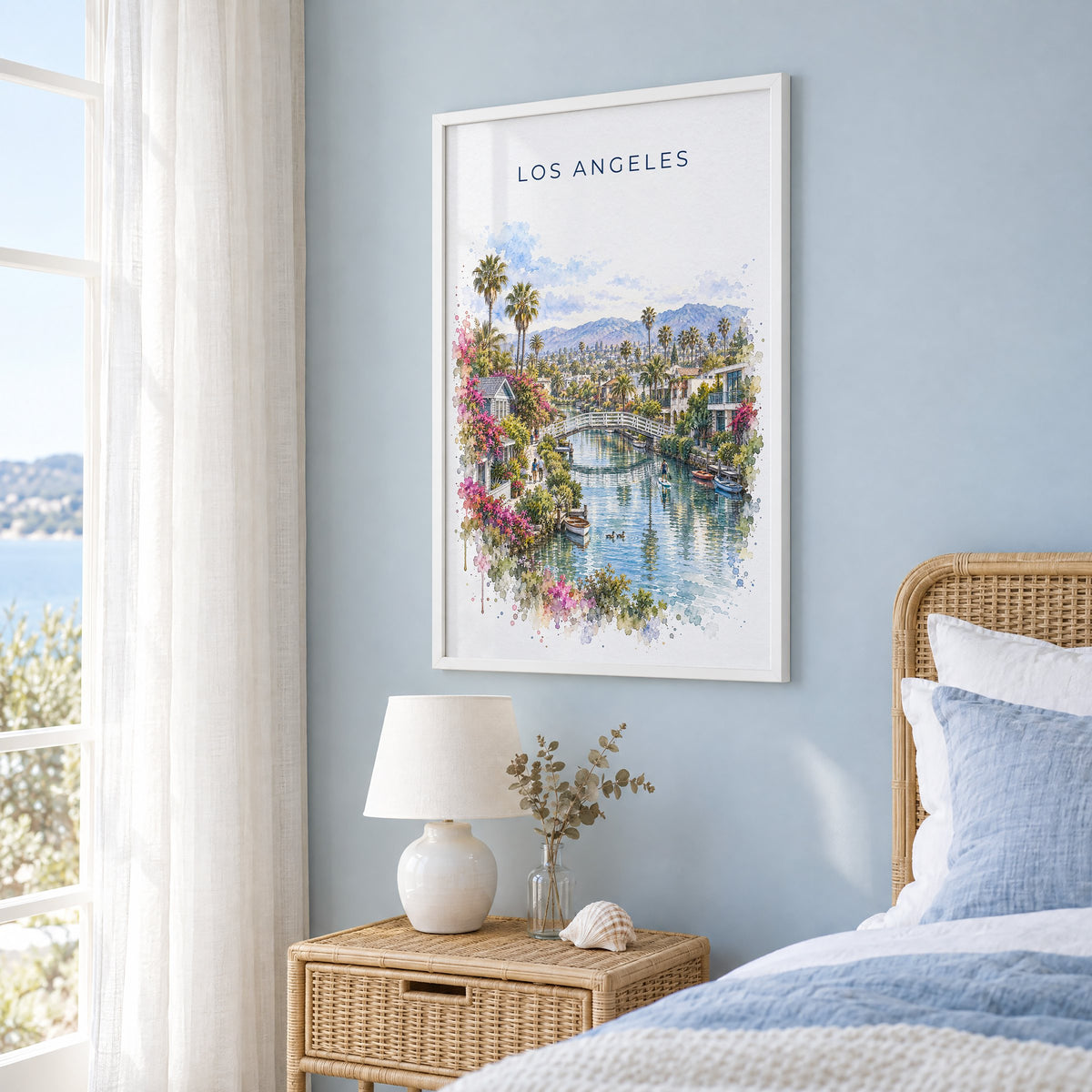 Venice Canals Los Angeles watercolor print in a white frame on a pale blue wall beside a rattan bed in a coastal bedroom