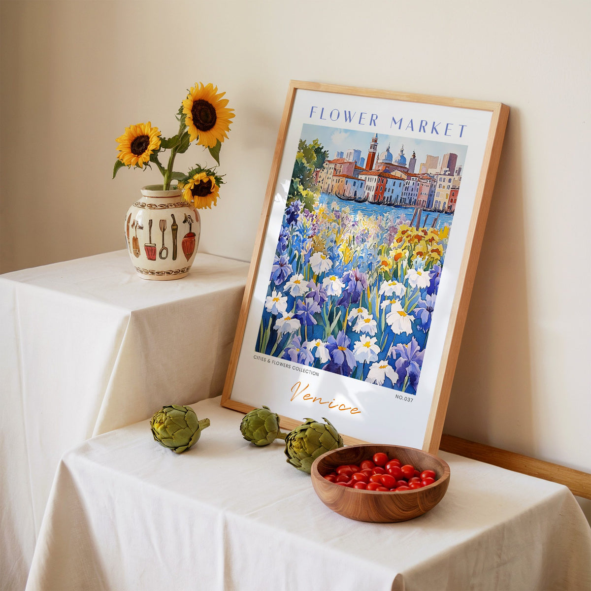 Venice flower market print in an oak frame leaning on a linen-draped table beside a vase of sunflowers