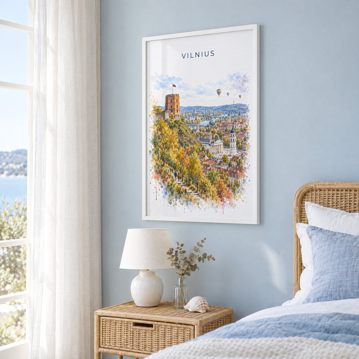 Vilnius watercolor print in a white frame on a pale blue wall beside a rattan bed in a coastal bedroom