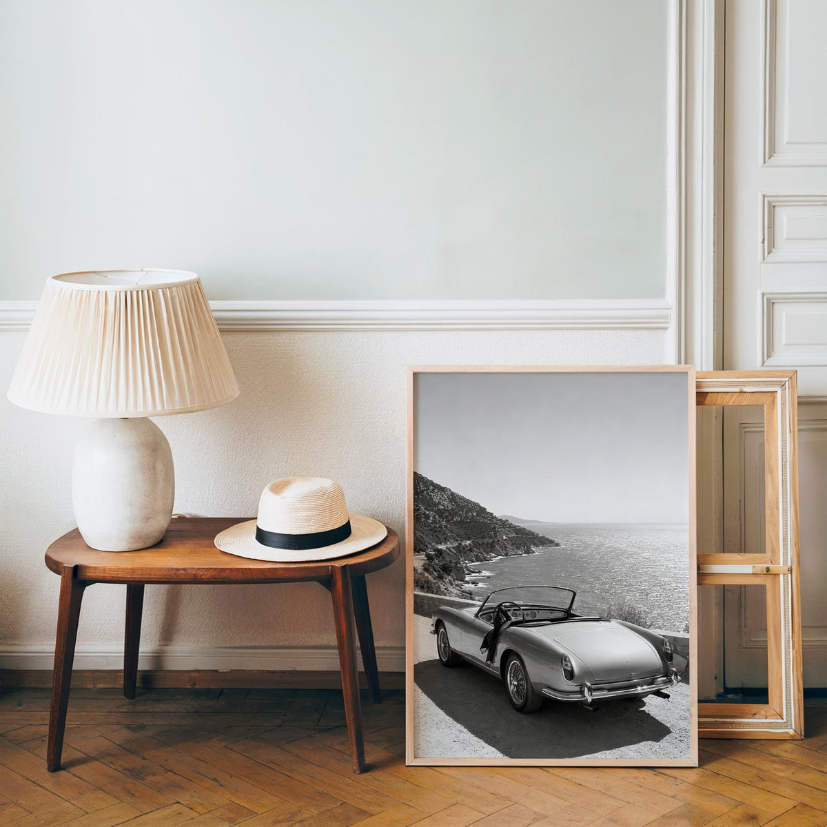 Vintage convertible parked above the sea print in a pine frame on a wood floor beside a lamp and straw hat