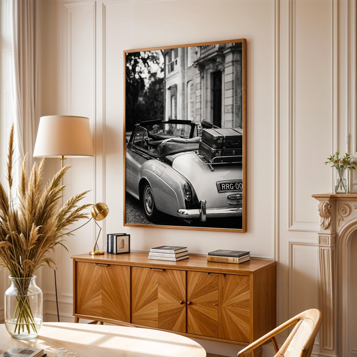 Woman in vintage convertible with leather suitcases print in an oak frame above a sunburst wood sideboard with pampas grass