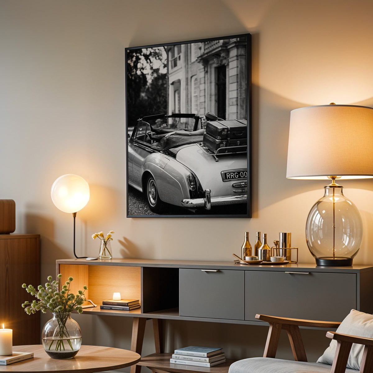 Woman in vintage convertible with leather suitcases print in a black frame above a grey media console with a glass table lamp