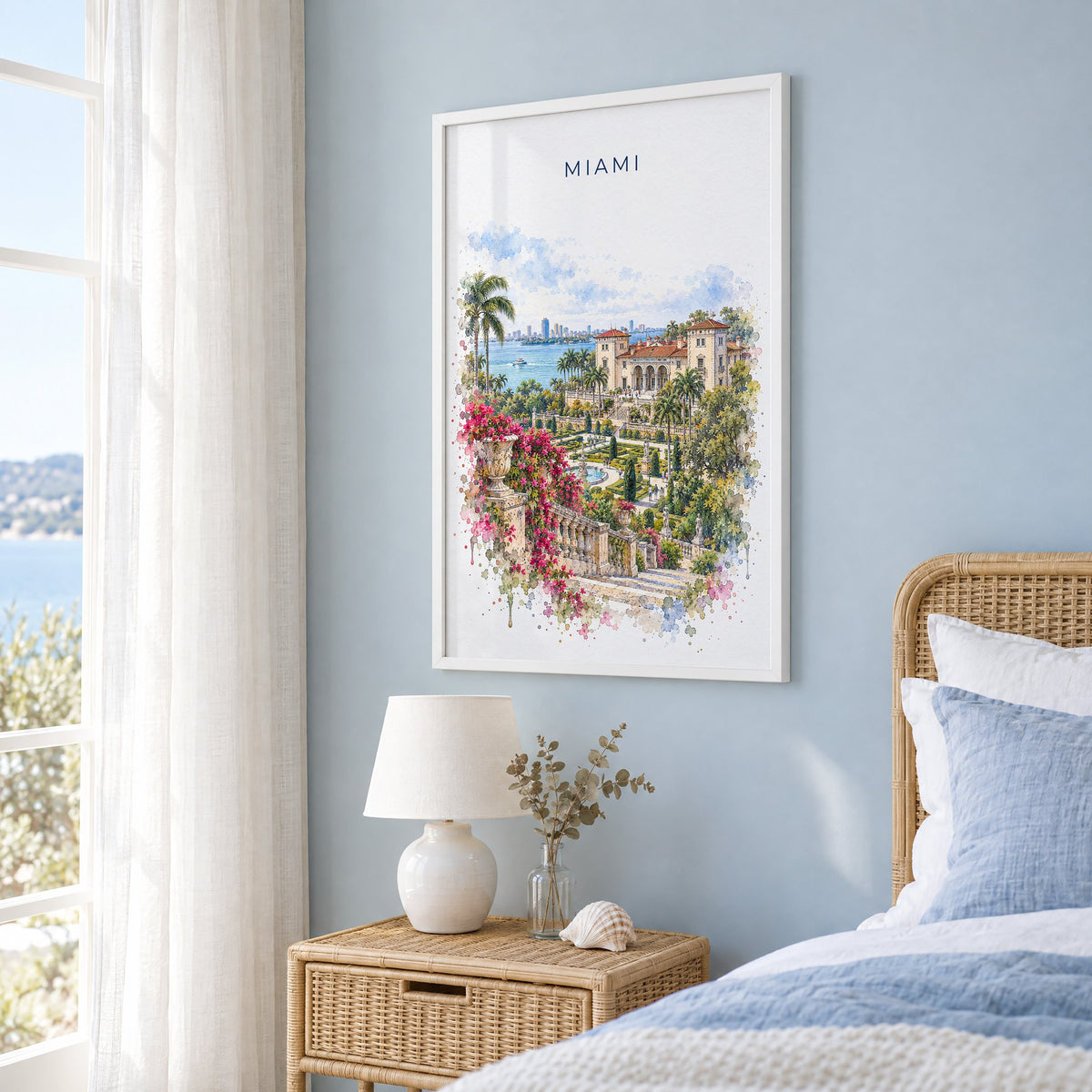 Vizcaya Miami watercolor print in a white frame on a pale blue wall beside a rattan bed in a coastal bedroom