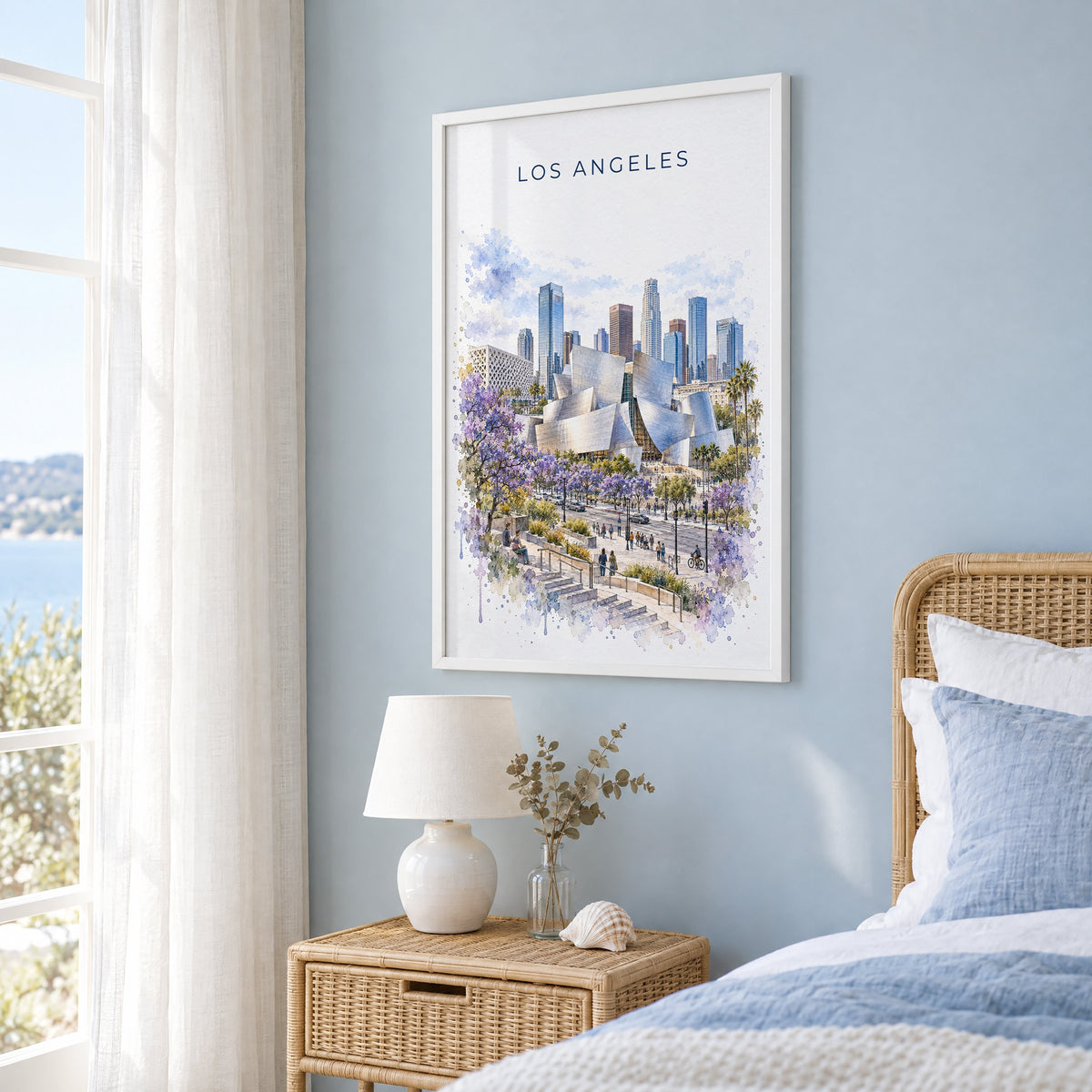 Walt Disney Concert Hall watercolor print in a white frame on a pale blue wall beside a rattan bed in a coastal bedroom