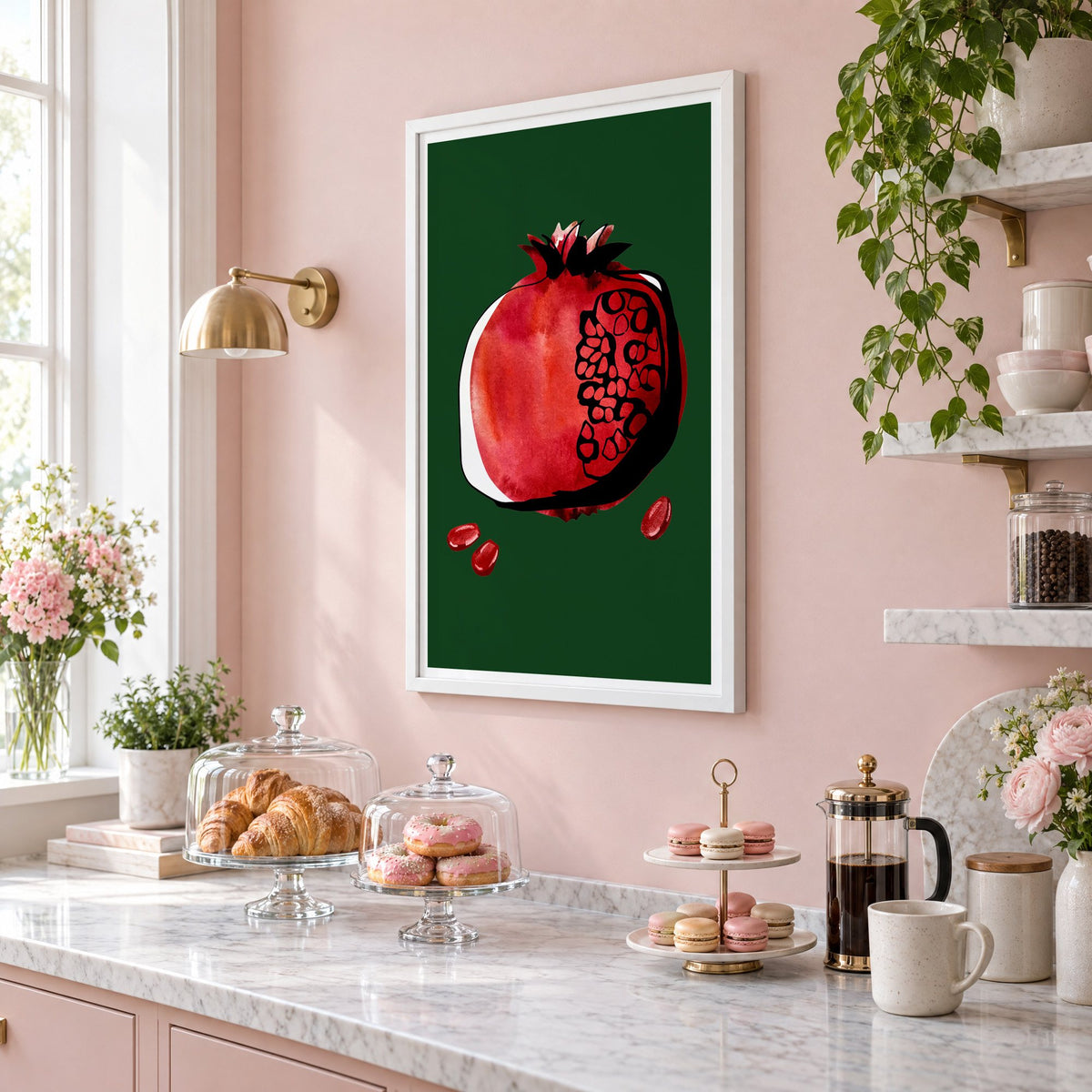 Red watercolor pomegranate on dark green print in a white frame on a blush pink wall above a patisserie counter with croissants and macarons, seen at an angle