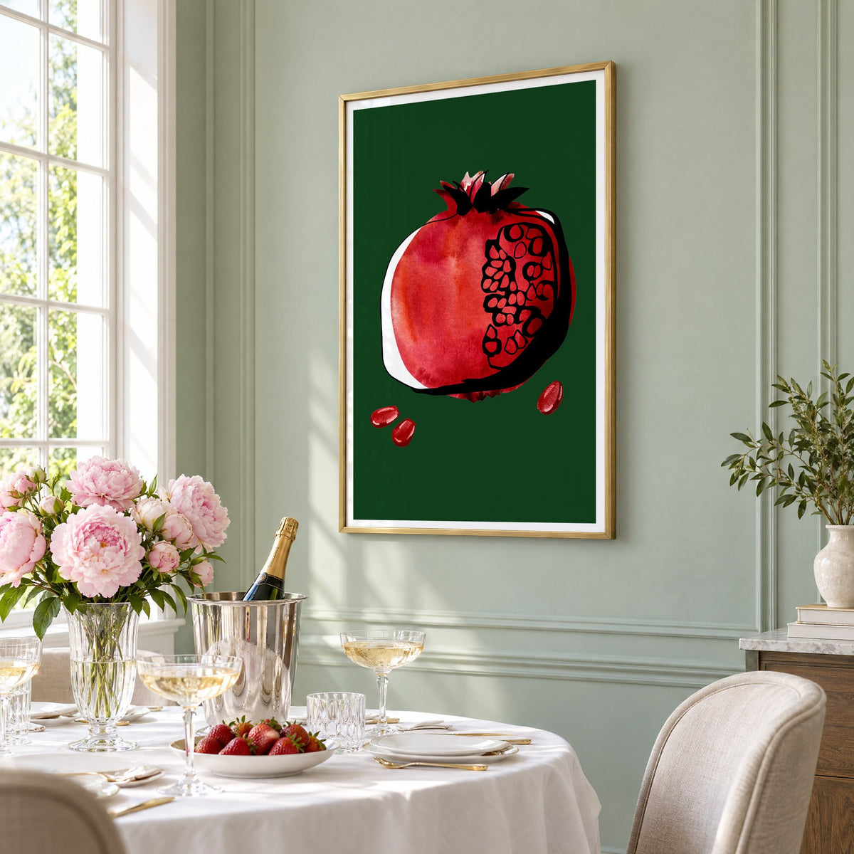 Red watercolor pomegranate on dark green print in a brass frame on a sage wall above a brunch table with champagne and peonies, seen at an angle