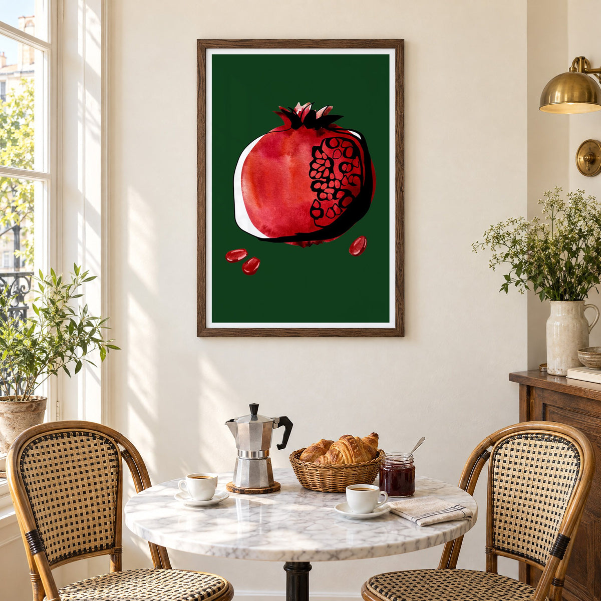 Red watercolor pomegranate on dark green print in a walnut frame above a marble bistro table with croissants and rattan chairs