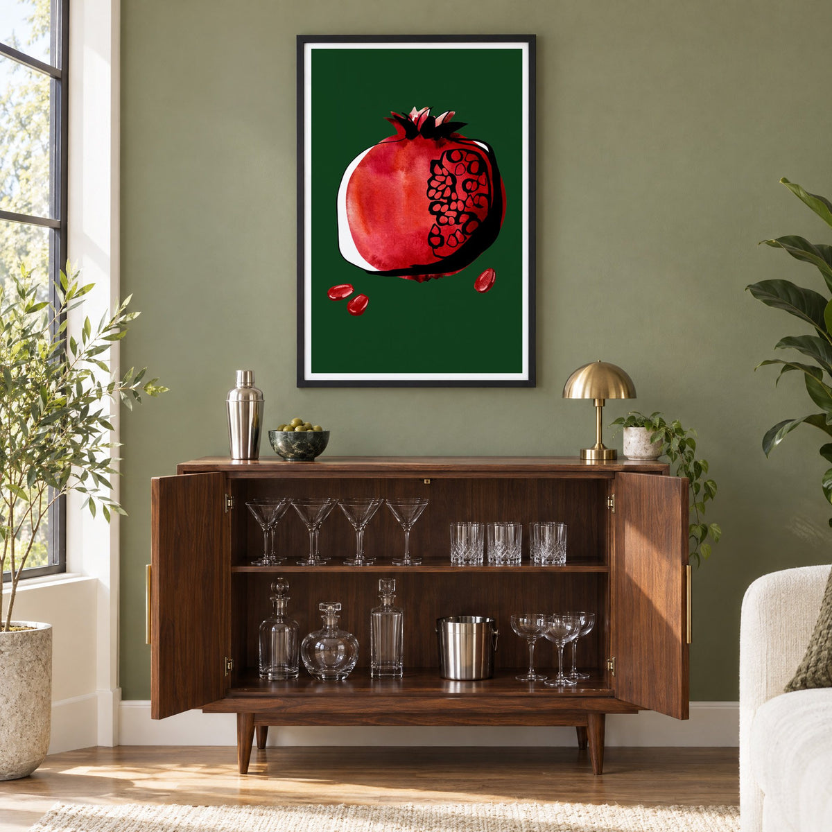 Red watercolor pomegranate on dark green print in a black frame on an olive green wall above a walnut bar cabinet with glassware