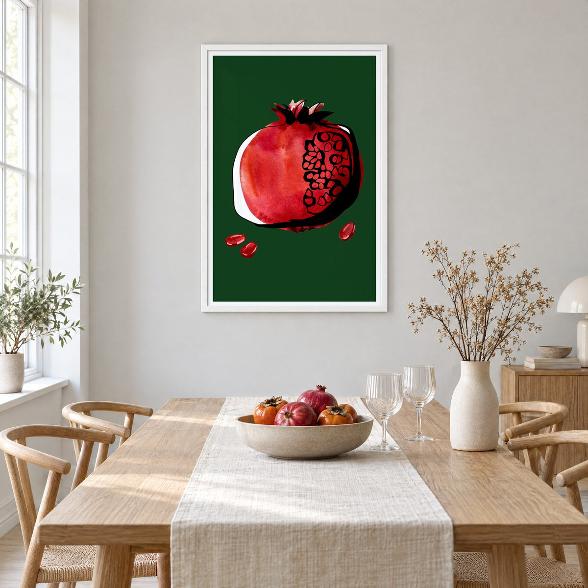 Red watercolor pomegranate on dark green print in a white frame above a light oak dining table with a linen runner and fruit bowl