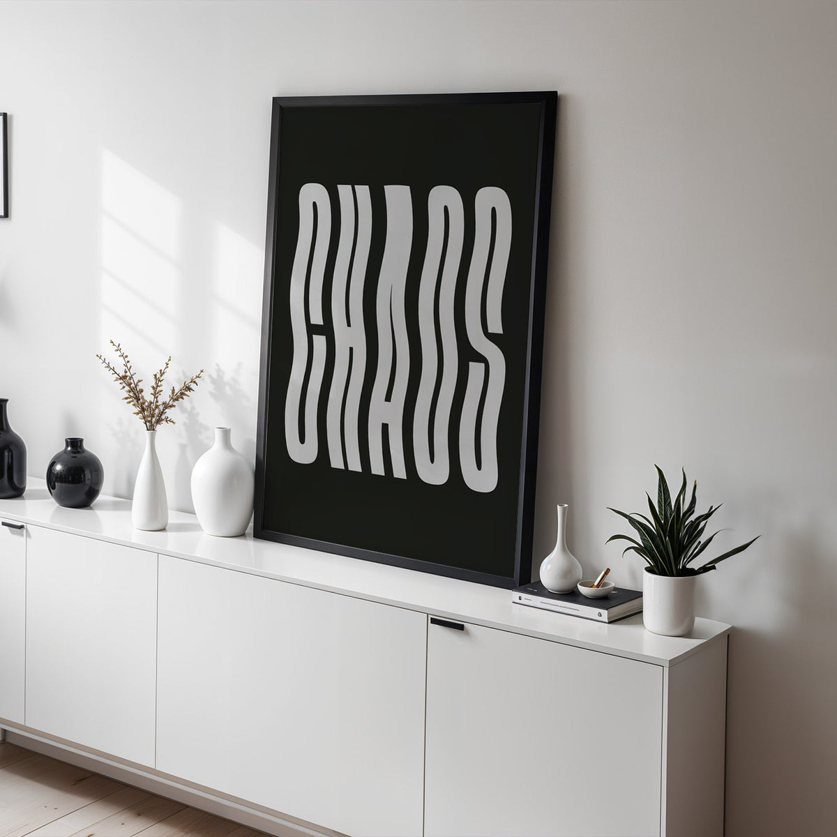Wavy light grey CHAOS lettering on black print in a black frame on a white sideboard with black and white vases