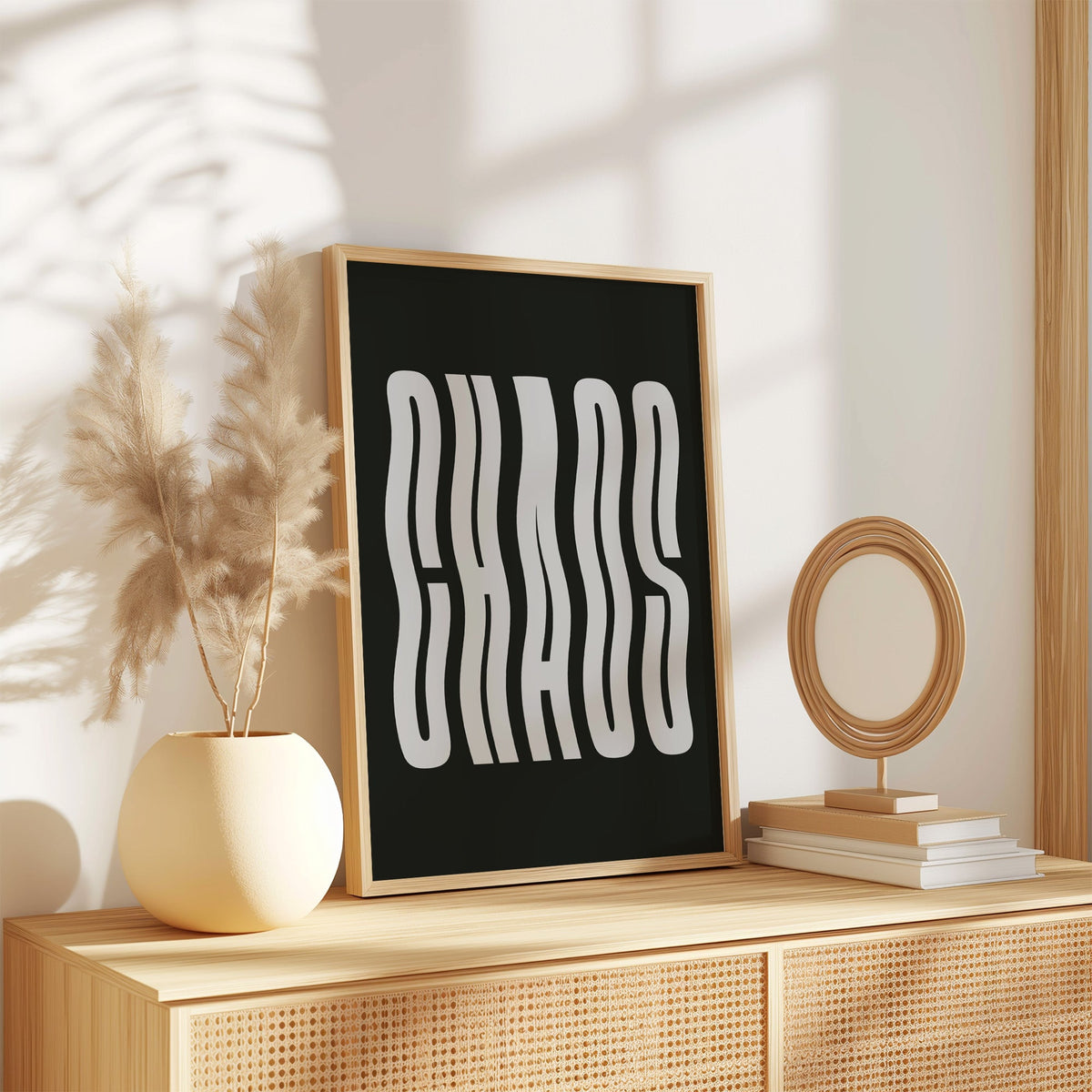 Wavy light grey CHAOS lettering on black print in an oak frame on a rattan sideboard beside pampas grass