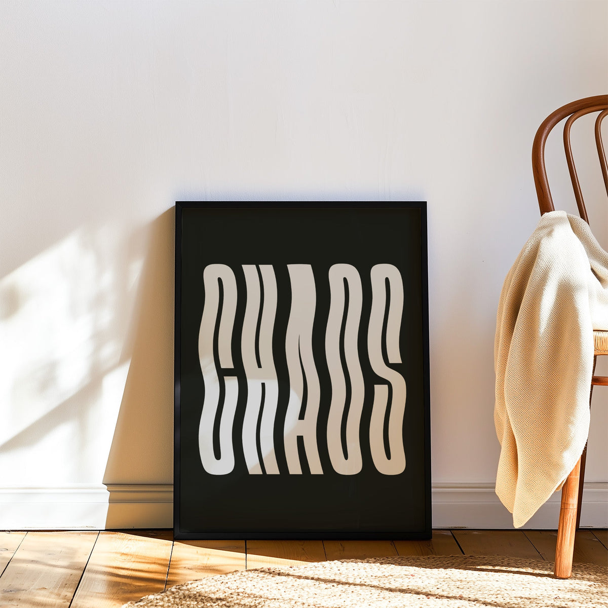 Wavy light grey CHAOS lettering on black print in a black frame leaning on a wood floor beside a bentwood chair