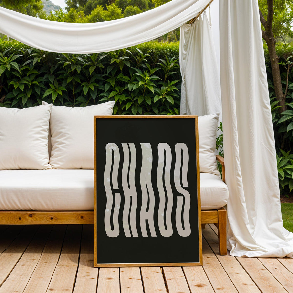 Wavy light grey CHAOS lettering on black print in an oak frame on an outdoor daybed under white drapes