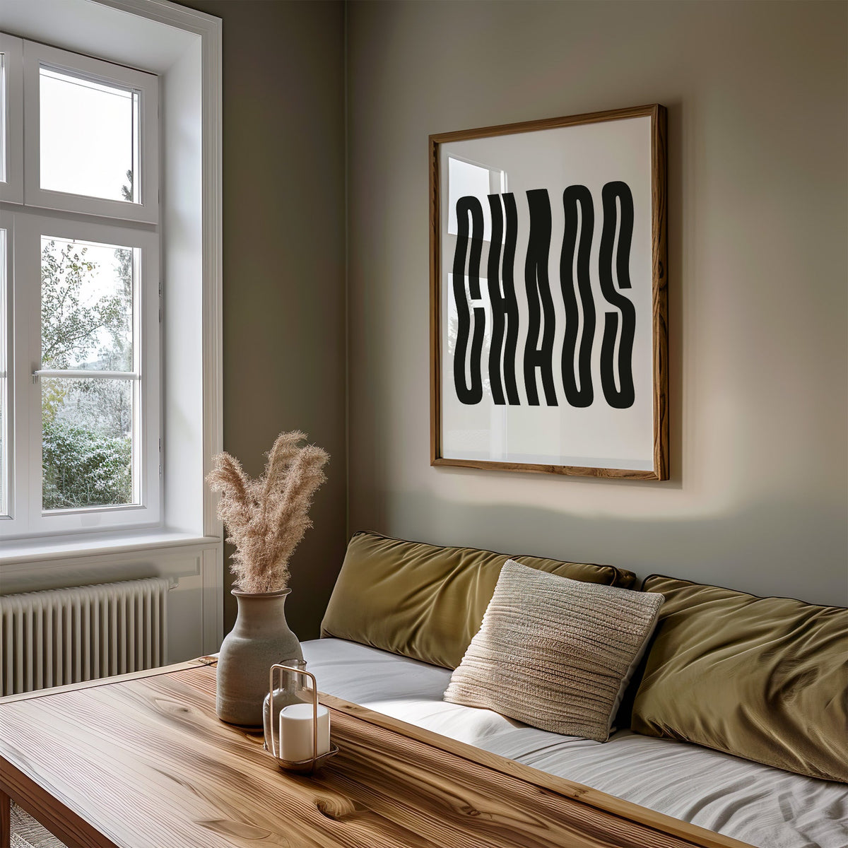 Wavy black CHAOS lettering on white print in a walnut frame above an olive daybed by a window