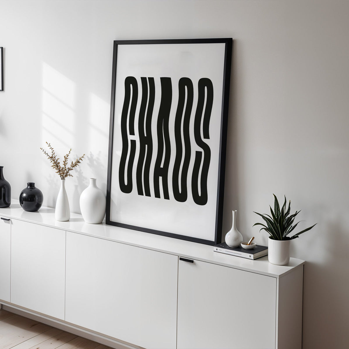 Wavy black CHAOS lettering on white print in a black frame on a white sideboard with black and white vases