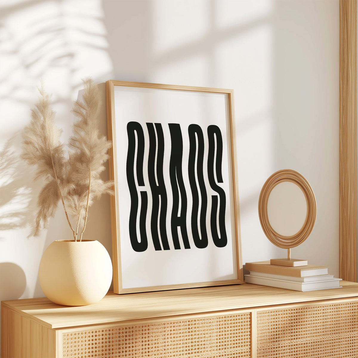 Wavy black CHAOS lettering on white print in an oak frame on a rattan sideboard beside pampas grass