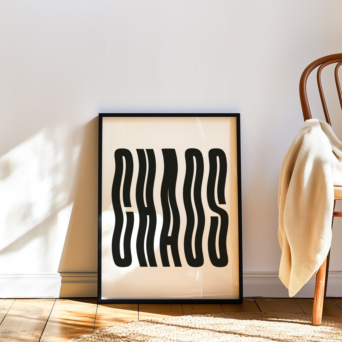 Wavy black CHAOS lettering on white print in a black frame leaning on a wood floor beside a bentwood chair