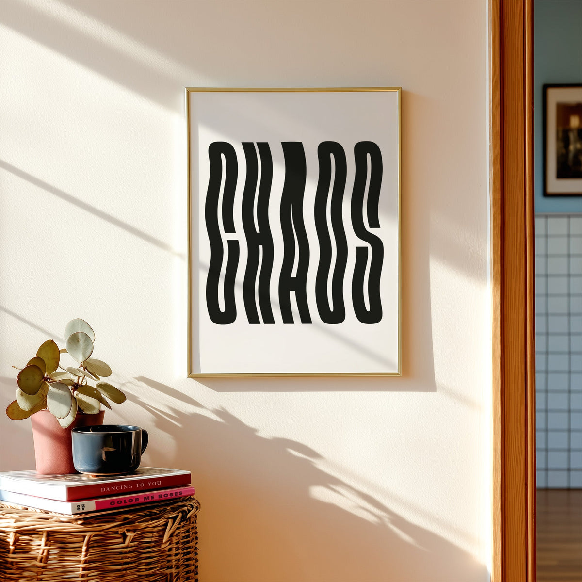 Wavy black CHAOS lettering on white print shown in a gold frame above a wicker basket and stacked books