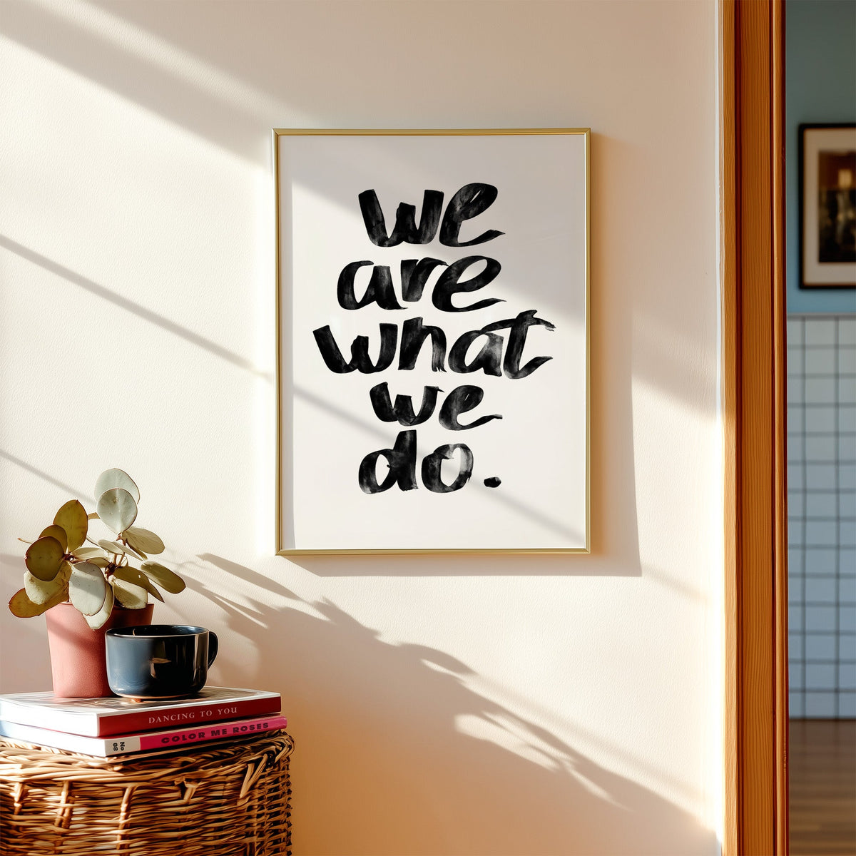 We are what we do brush lettering quote print shown in a gold frame above a wicker basket and stacked books