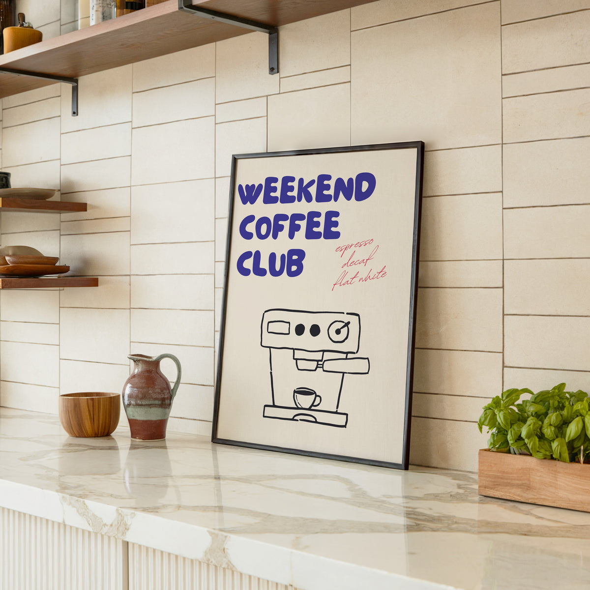 Weekend Coffee Club print in a black frame leaning on a marble counter in a tiled kitchen