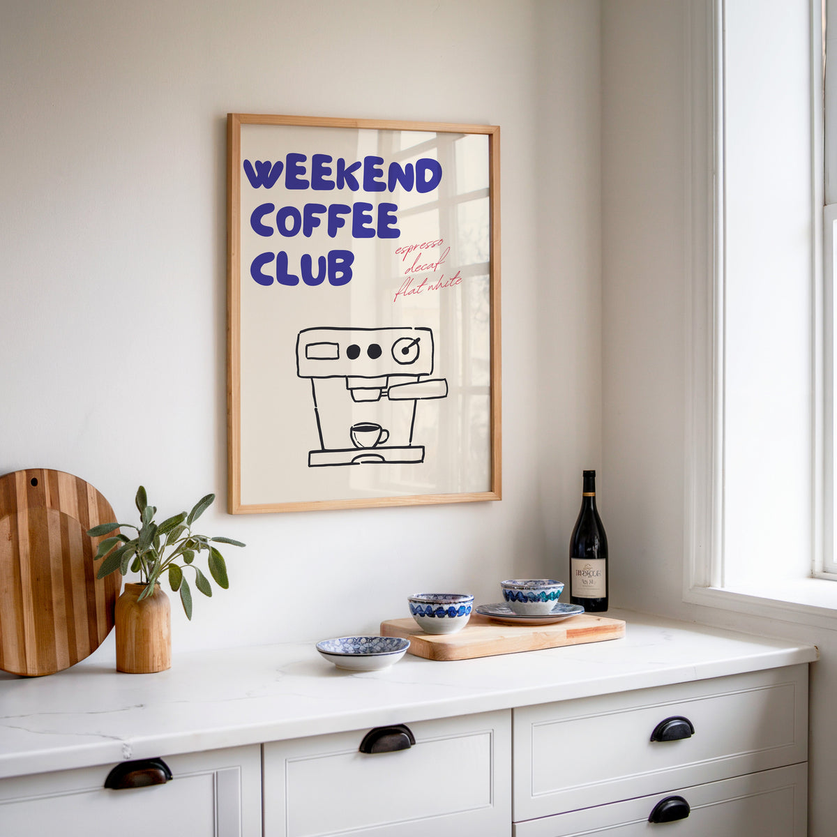 Weekend Coffee Club print in an oak frame above a white sideboard with a bottle of wine