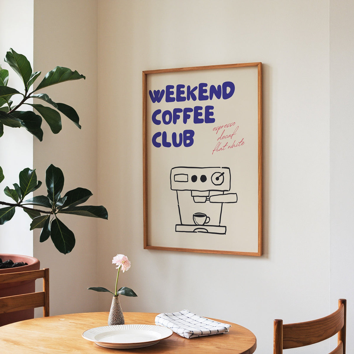 Weekend Coffee Club print in a walnut frame above a round table beside a fiddle-leaf fig