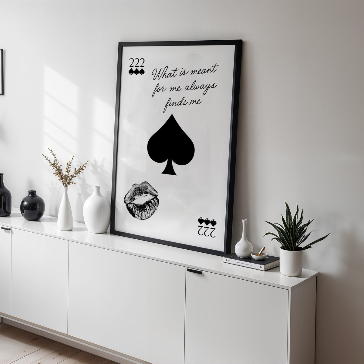 Spade, kiss mark and script quote print in a black frame on a white sideboard with black and white vases