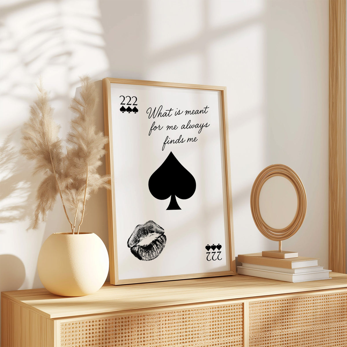 Spade, kiss mark and script quote print in an oak frame on a rattan sideboard beside pampas grass
