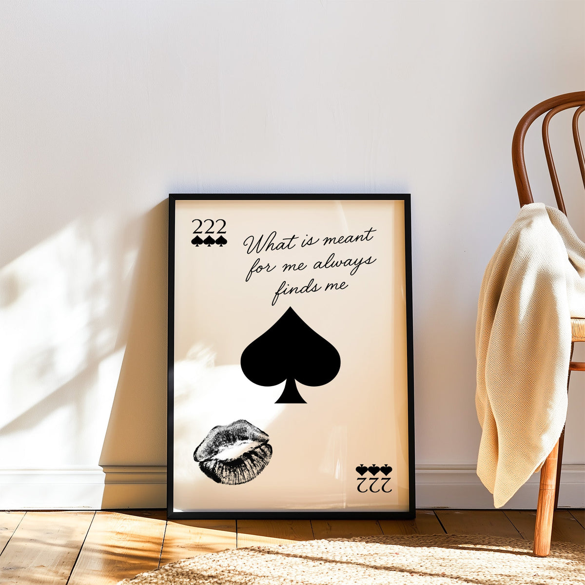 Spade, kiss mark and script quote print in a black frame leaning on a wood floor beside a bentwood chair