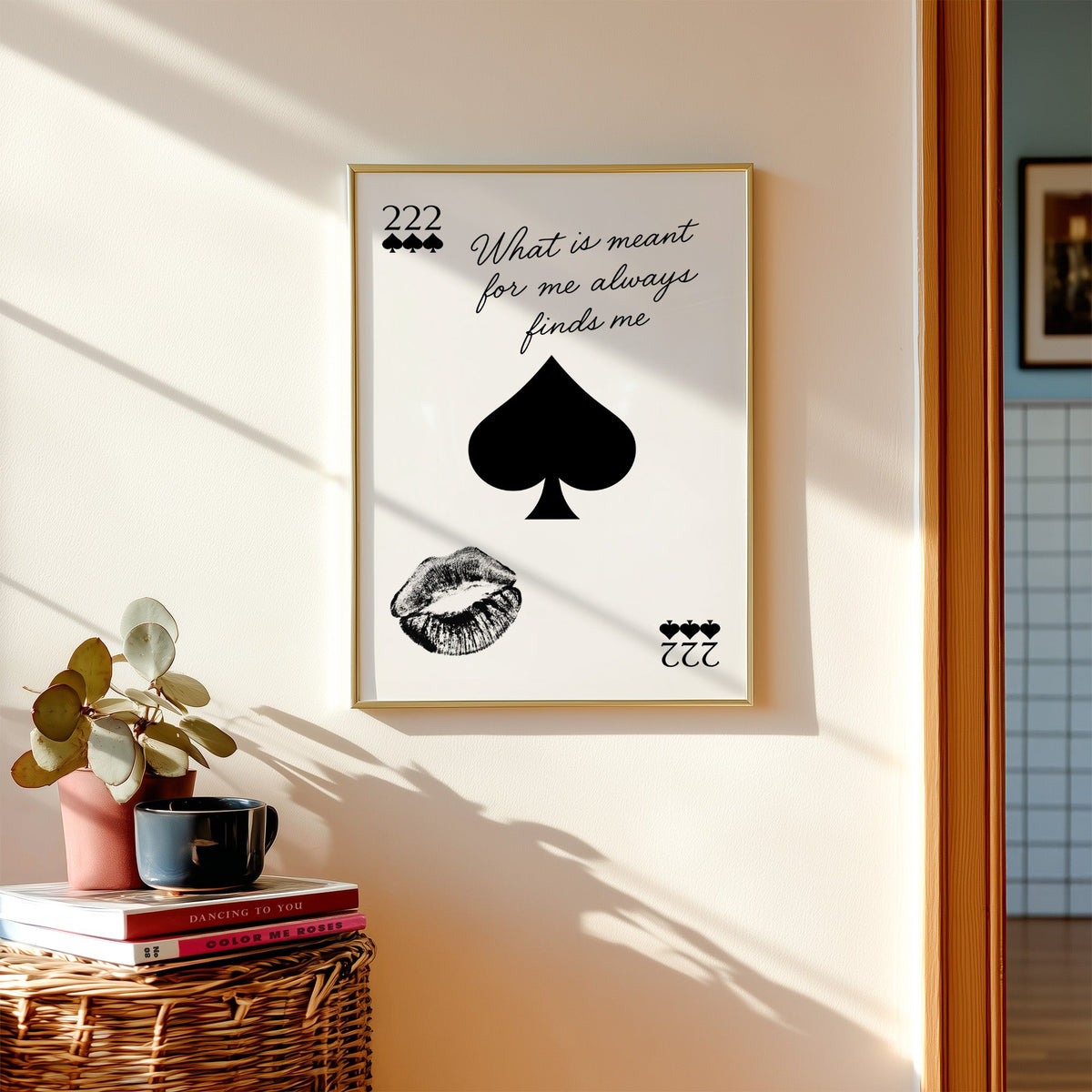 Spade, kiss mark and script quote print shown in a gold frame above a wicker basket and stacked books